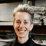 A white woman smiling at the camera. The photo is cropped at the top of her shoulders. She is wearing a black jacket, long earrings, and has short hair dyed platinum blonde on top and shaved shorter on the sides.