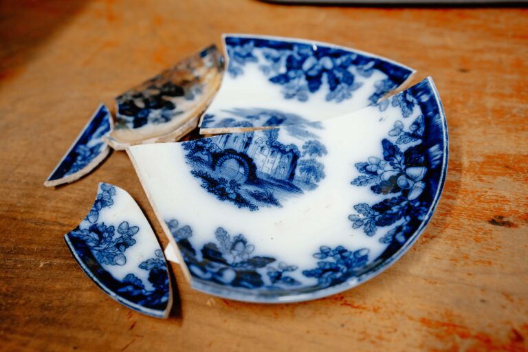 The broken pieces of an unearthed ceramic plate. It is white with deep blue floral art painted around the outside with what appears to be a building painted in the center. The pieces rest on a worn wooden table.